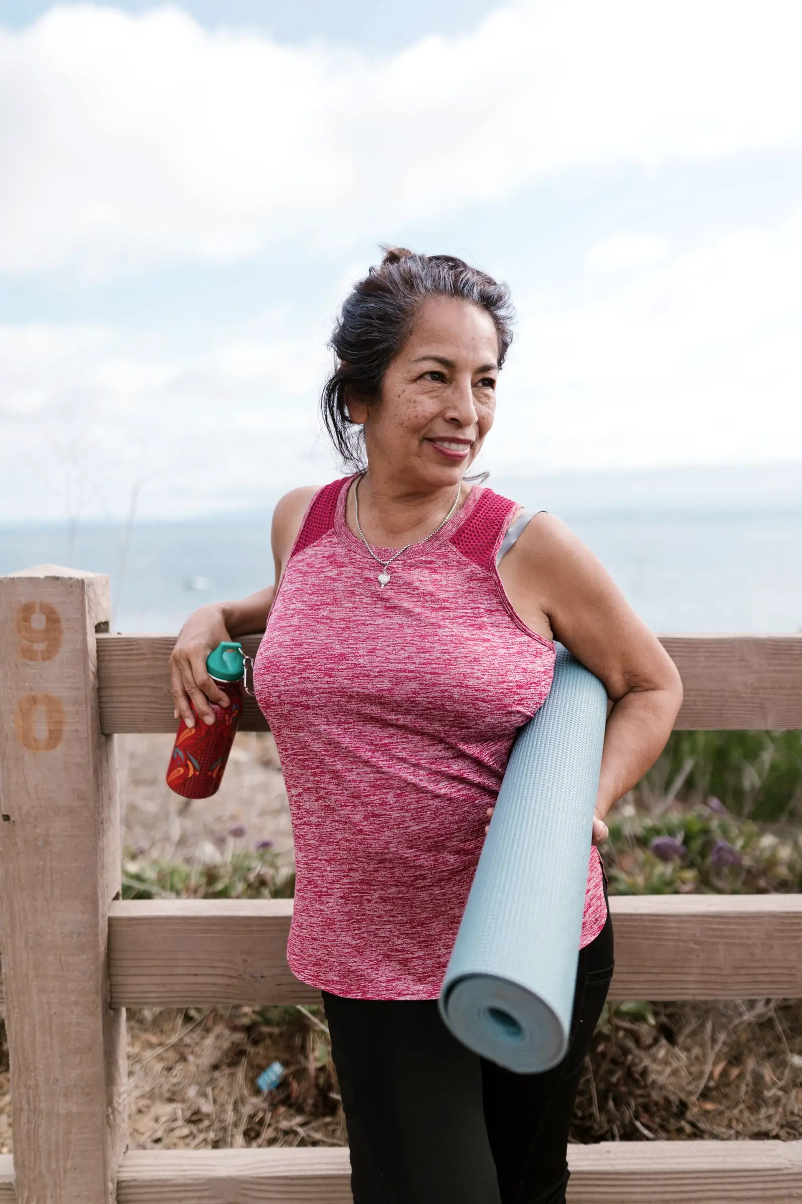 A woman over 60 carrying a yoga mat by the sea